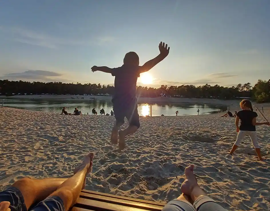 Child jumping at the sandy beach in the evening sun in front of the lake at Südsee-Camp; in the foreground, relaxed feet of two people on a bench