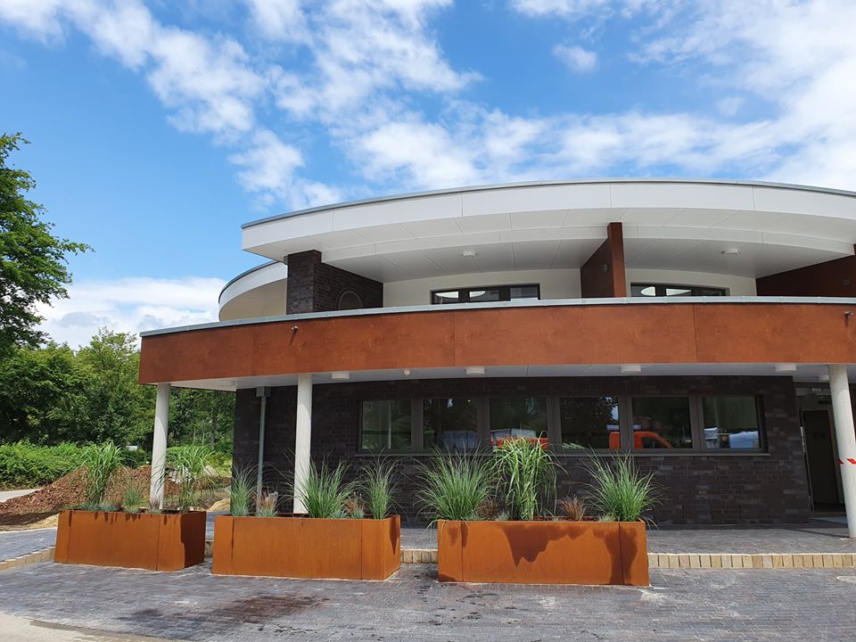 Learn more Modern round-shaped sanitary building with large windows and rust-colored facade elements. Planters with ornamental grasses are placed in front of the building.