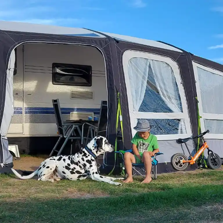 Junge sitzt barfuß vor einem Vorzelt am Südsee-Camp neben einem Dalmatiner-Hund, im Hintergrund ein Wohnwagen