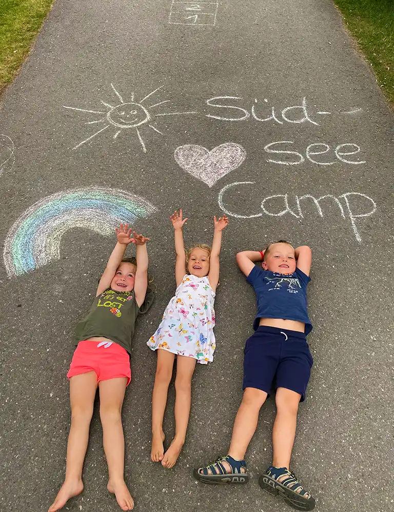 Drei Kinder liegen barfuß lachend auf einem Asphaltweg, auf dem mit bunter Kreide eine Sonne, ein Herz, ein Regenbogen und der Schriftzug „Südsee-Camp“ gemalt wurden.