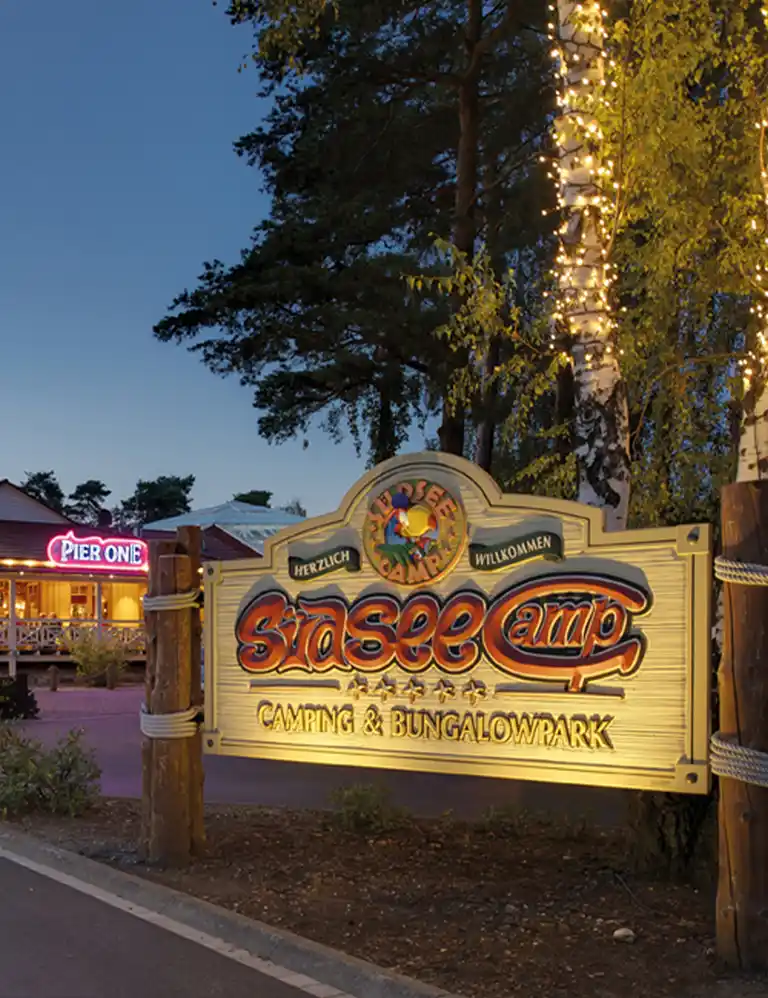 Einfahrt zum Südsee-Camp bei Abenddämmerung mit beleuchtetem Willkommensschild und Lichterketten an den Bäumen.