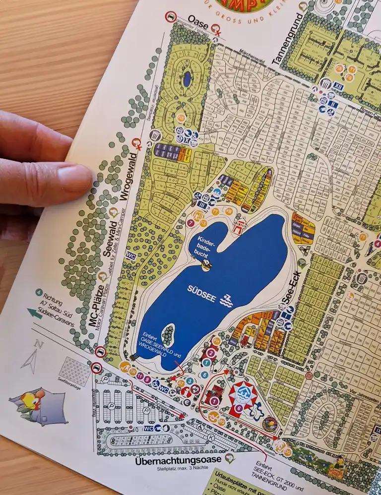 Übersichtskarte des Südsee-Camps mit eingezeichneten Bereichen wie dem Südsee-See, Campingplätzen, Sanitärgebäuden, Spielplätzen und thematisch benannten Arealen wie „Seewald“, „Wrogewald“ und „Oase“. Eine Hand hält die Karte am Rand fest.