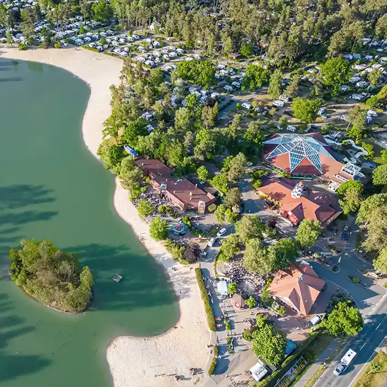view other areas Aerial view of Südsee-Camp featuring the central swimming pool with a distinctive glass roof. Surrounded by trees, camping pitches, and a large sandy beach lake. The well-maintained resort offers various recreational facilities in a natural setting.