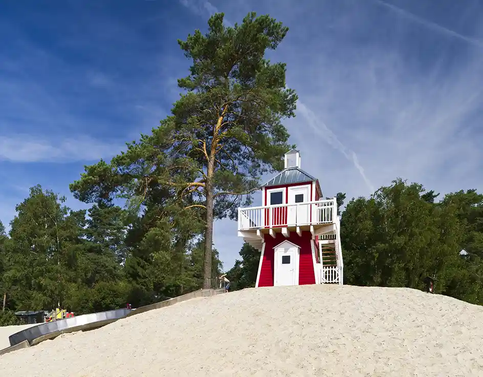Preise 2025 & 2026: Ferienhaus, Chalet, Mietwohnwagen | Südsee-Camp Roter Leuchtturm mit weißem Balkon am Strand des Südsee-Camps, daneben eine lange Rutsche und umgeben von Kiefern unter blauem Himmel