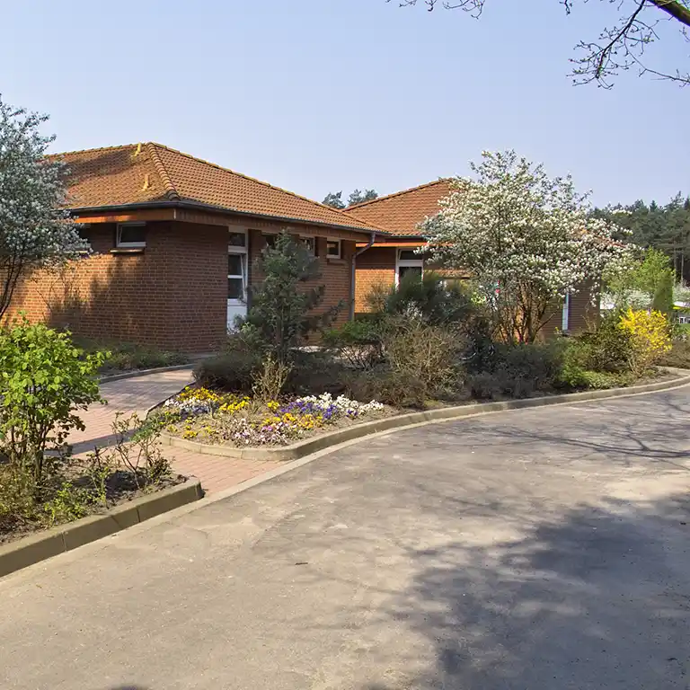 Learn more Sanitary building 10 at Südsee-Camp – a red-brick facility with a tiled roof, surrounded by well-tended flower beds and blooming trees, nestled in a natural, forest-adjacent setting.