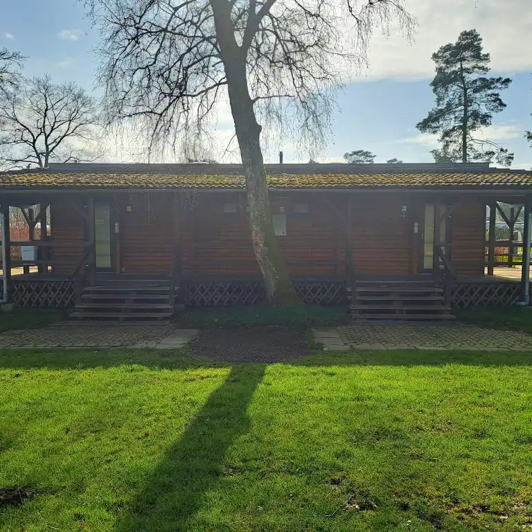 Learn more Wooden sanitary building in the Tannengrund area of Südsee-Camp, featuring two separate entrances and a covered porch, surrounded by grass and trees – campsite toilet facilities for guests with dogs.