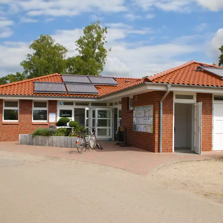 Learn more Sanitary building 2 at Südsee-Camp featuring a red-tiled roof, solar panels, and a neatly landscaped entrance with a bicycle parking area – clean facilities under a sunny sky.