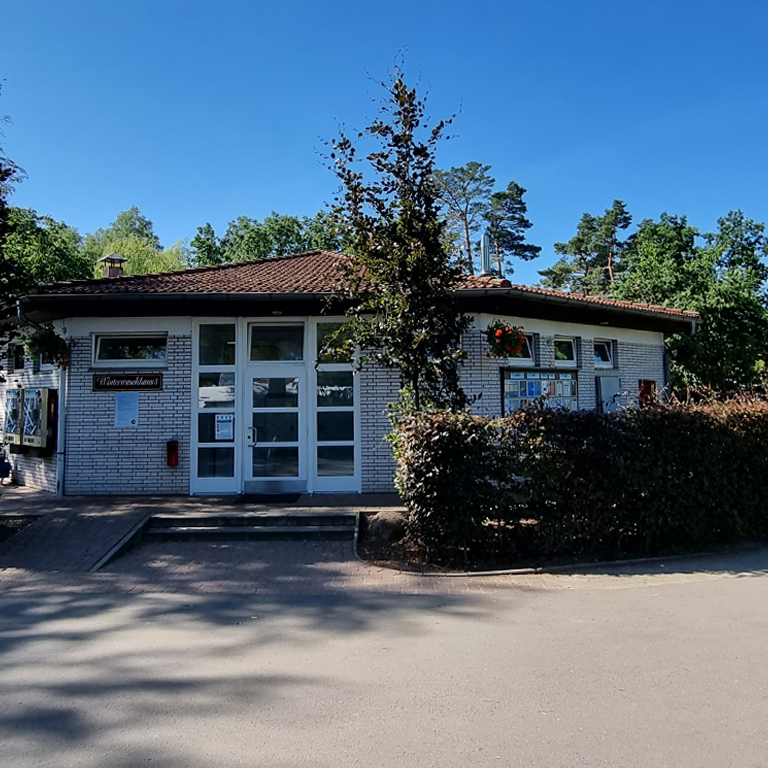 Learn more Sanitary building 5 at Südsee-Camp with light-colored brick walls, a brown tiled roof, flower decorations on the windows, and surrounded by trees and hedges.