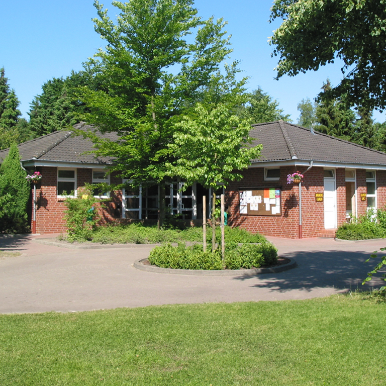 Learn more Washhouse 6 at Südsee-Camp, a red brick building with a dark hip roof, well-kept front garden and bulletin boards, surrounded by trees and greenery on a sunny summer day.