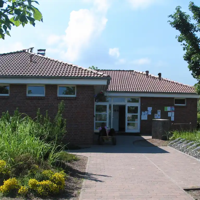 Learn more Exterior view of sanitary building 7 at Südsee-Camp with red tiled roof, brick facade, accessible walkway, flower beds, and bicycle racks – a modern, well-maintained sanitary facility in a green setting