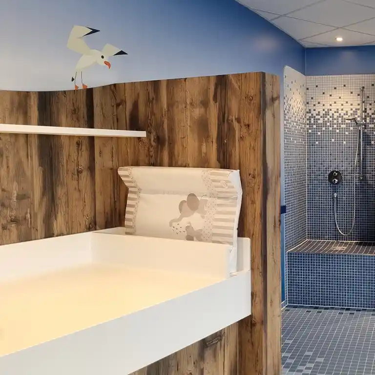 Changing table in the children’s bathroom of sanitary building 4 at Südsee-Camp, featuring a cushioned pad, wooden paneling, and a tiled shower area in the background. The wall is painted blue with a seagull illustration.