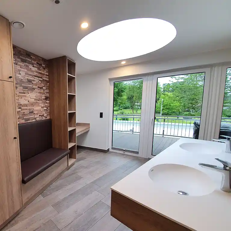 Modern private bathroom in sanitary building 4 at Südsee-Camp featuring two sinks, a wooden bench, open shelves, a make-up table, and a large round skylight that lets in natural light. Glass doors open to a terrace with a view of the greenery.