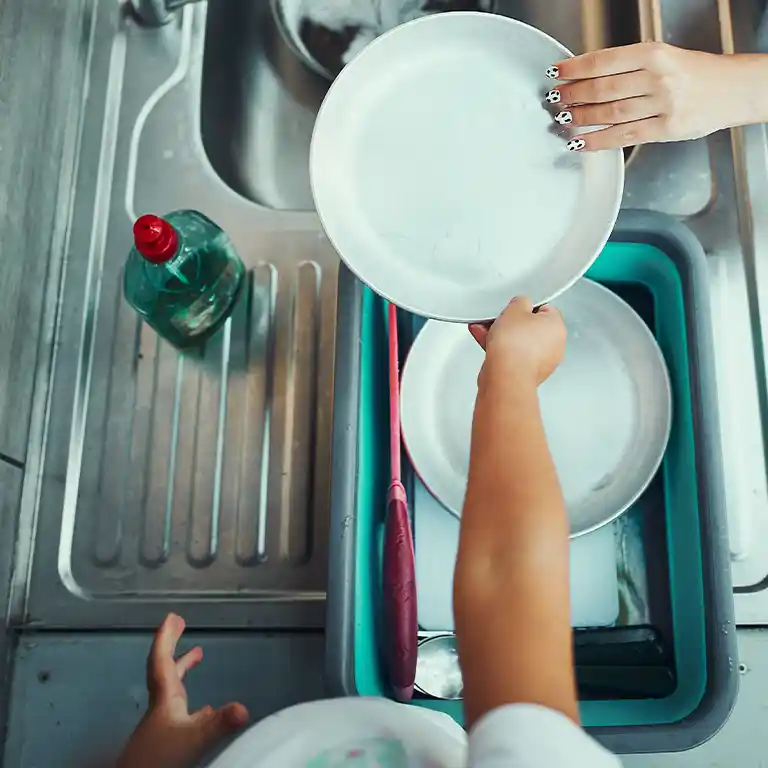 [Translate to English:] Zwei PersTwo people are washing dishes together in the dishwashing area of the South Sea Camp. Visible are a stainless steel sink with draining board, a green washing tub filled with plates, and a bottle of dish soap. The focus is on handing over a clean plate.onen spülen gemeinsam Geschirr in einem Spülraum des Südsee-Camps. Zu sehen ist eine Edelstahlspüle mit Abtropffläche, eine grüne Spülschüssel mit Tellern und eine Flasche Spülmittel. Der Fokus liegt auf dem Hantieren mit sauberen Tellern.