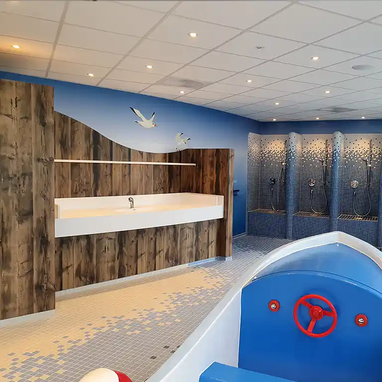Child-friendly shower and wash area with maritime theme at sanitary building 4 of Südsee-Camp. In the foreground, a blue toy boat with a red steering wheel; in the background, several kid-sized showers with blue mosaic tiles and a sink in front of a wood-style wall decorated with seagulls.