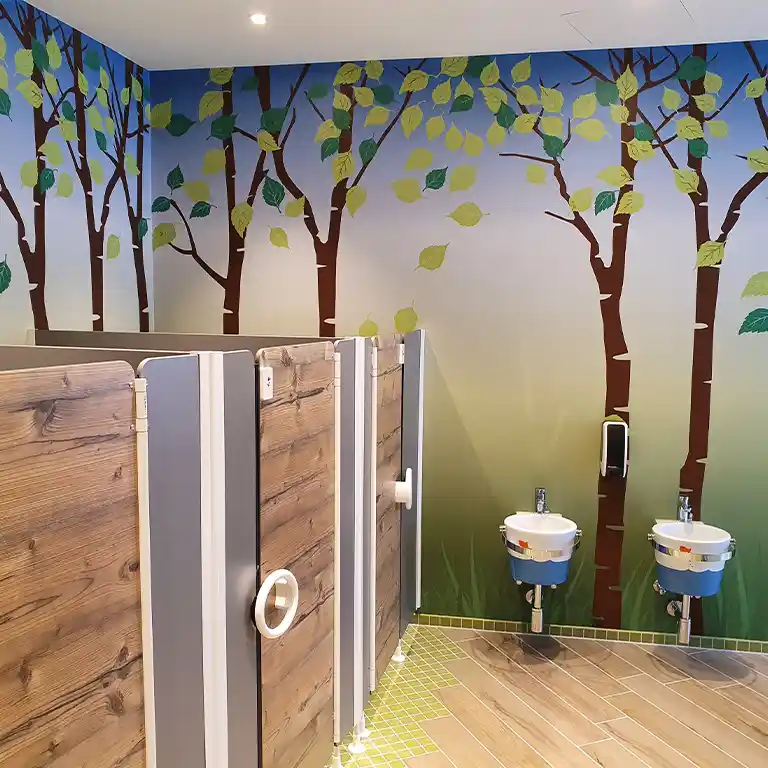 Children’s sanitary area at sanitary building 4 of Südsee-Camp with a playful forest-themed wall design. Child-sized toilet cubicles in a wood look and colorful small sinks create a cheerful, family-friendly environment.
