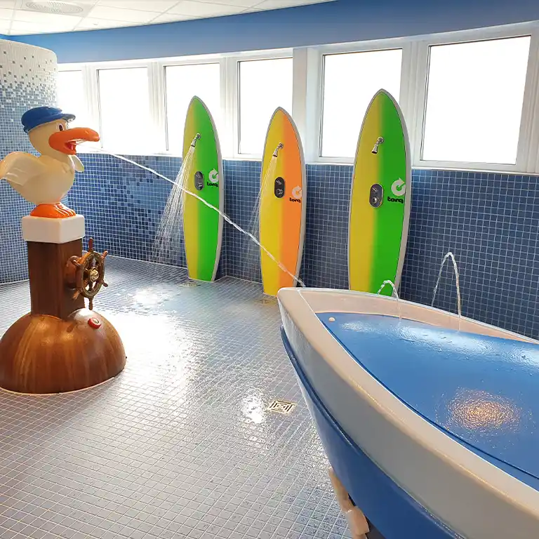 Playful children’s washroom in sanitary building 4 at Südsee-Camp with blue mosaic tiles, colorful surfboard showers, and a seagull figure spraying water. A boat-shaped play structure with fountains is in the foreground.