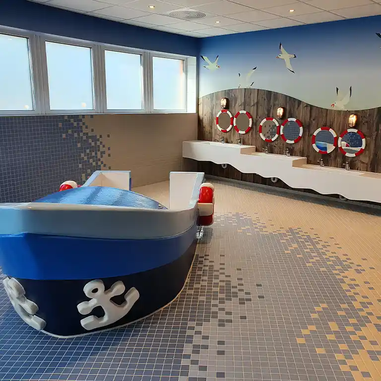 Maritime-themed children’s washroom at sanitary building 4 of Südsee-Camp, featuring a playful blue-and-white toy boat, child-friendly sinks, and mirrors framed as life rings. Seagulls painted on the wall create a cheerful harbor scene.