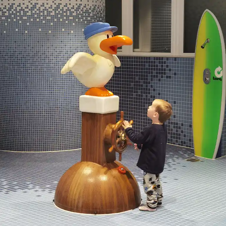 Toddler playing in the children’s washroom at sanitary building 4 in Südsee-Camp, turning a ship’s wheel attached to a seagull figure wearing a sailor’s cap. In the background: colorful surfboard showers and blue tiled walls.