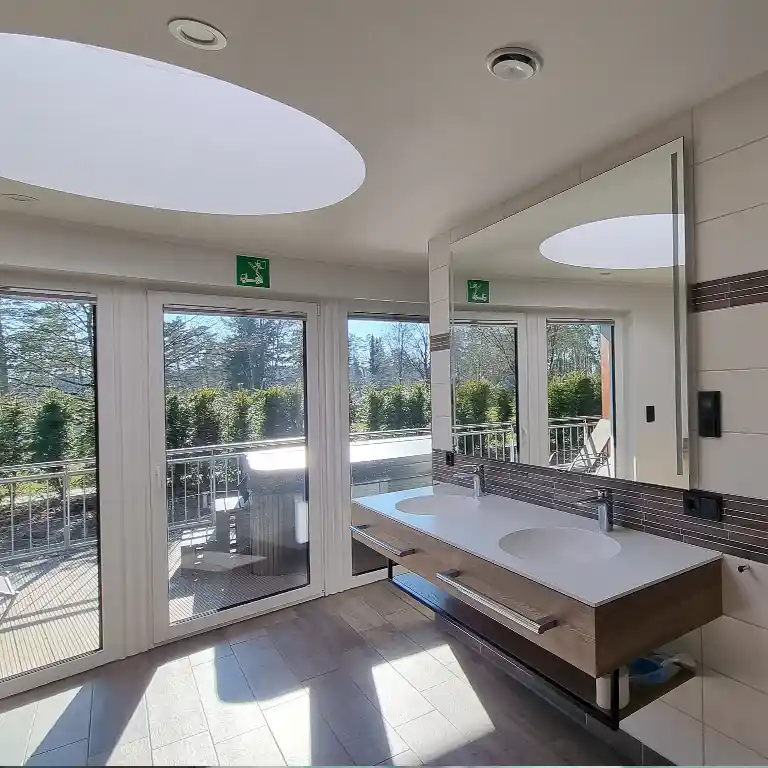 Modern private bathroom in sanitary building 4 at Südsee-Camp with two sinks, a large mirror, natural daylight from a circular skylight, and access to a private terrace with garden view.