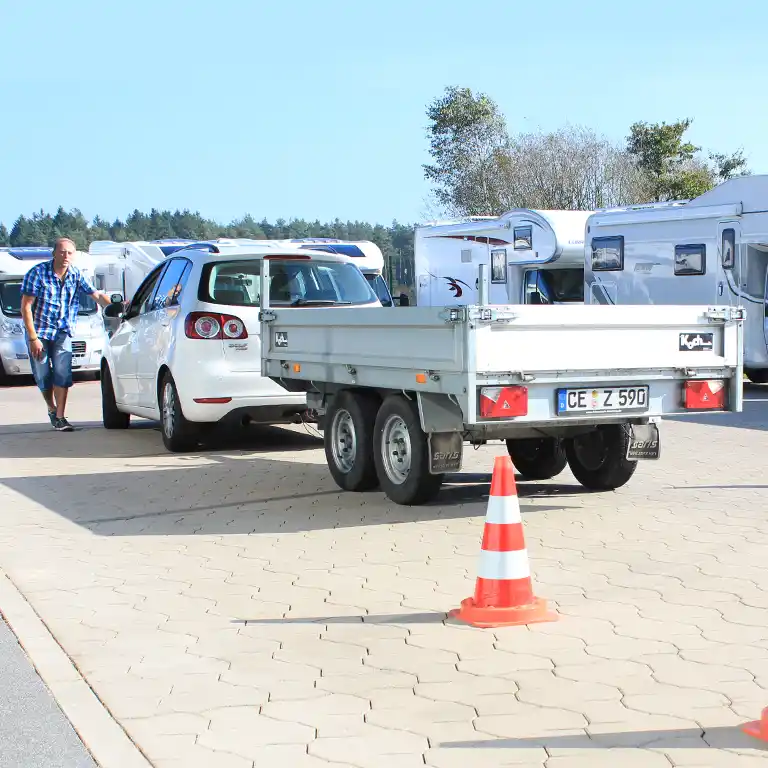 A participant of the B96 training course practices maneuvering with a trailer at the Südsee-Camp site. A white car with a trailer stands between traffic cones, with caravans and campers in the background. Practical driving training for safe travel with trailers and caravans.