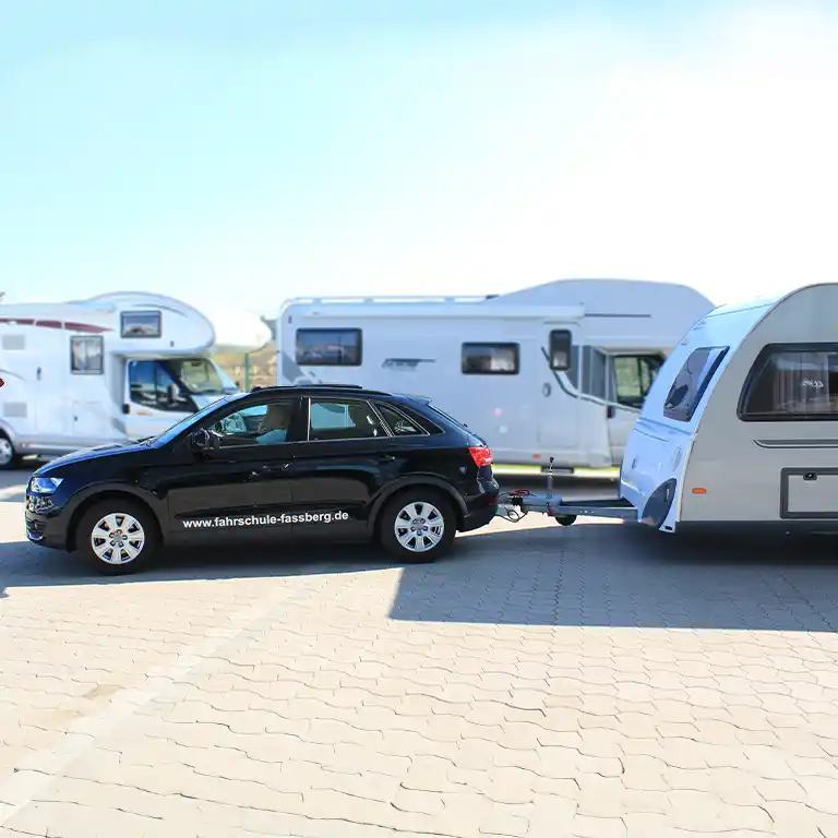 A black driving school car tows a caravan trailer on the Südsee-Camp grounds. Several motorhomes are visible in the background. During the B96 course, participants learn safe driving with a trailer – perfect for future campers and caravan travelers.