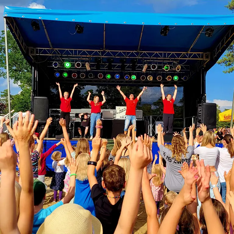 A joyful crowd of families and children dances and raises their hands in front of a stage at Südsee-Camp. On stage, four young entertainers in red shirts lead the audience under sunny skies. Summery atmosphere full of fun and togetherness at the family event in Südsee-Camp.