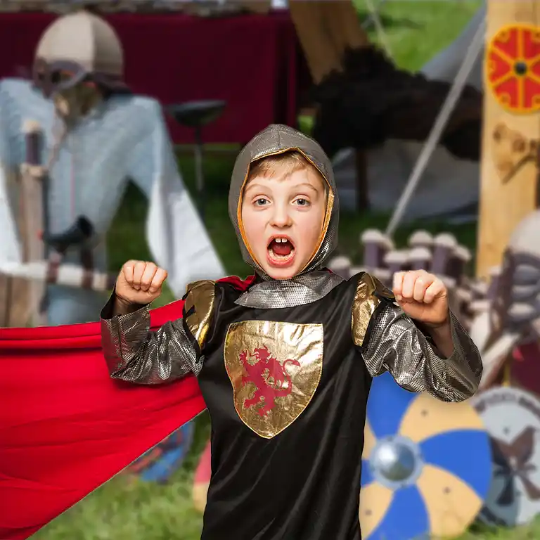 Ein Kind in einem Ritterkostüm mit goldenem Wappen und rotem Umhang spielt begeistert auf einem Mittelalterfest im Südsee-Camp. Im Hintergrund sind Helme, Schilde und Zelte zu sehen. Ein fantasievolles Erlebnis für kleine Abenteurer und Familien.