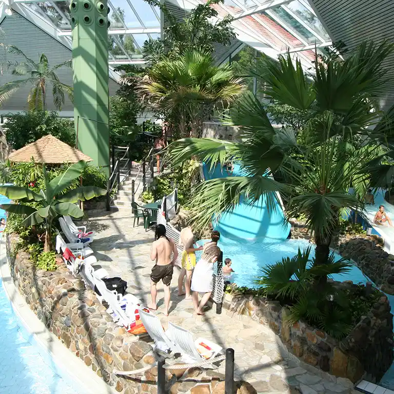 Tropischer Innenbereich im Südsee-Camp mit Wasserrutsche, Palmen, Liegestühlen und Badegästen im Lagunenpool unter einem Glasdach.