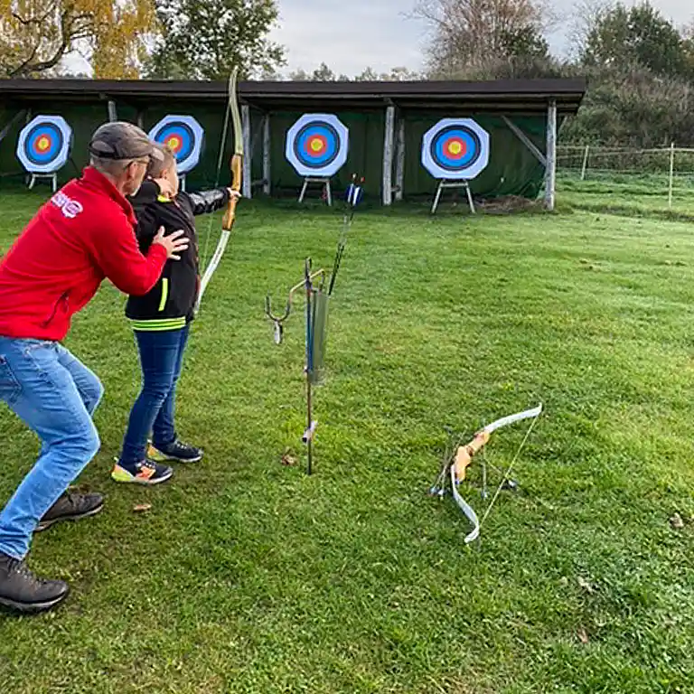 An entertainer assists a child with archery on a green lawn with targets in the background. The child holds the bow drawn while the entertainer helps adjust the posture.