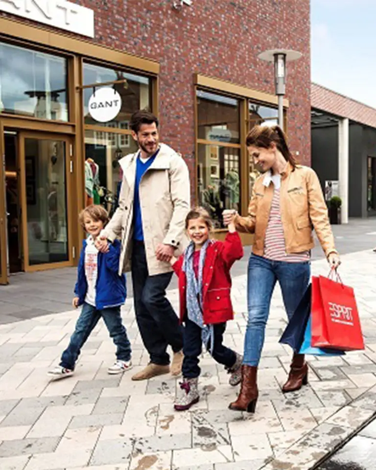 zum Shopping-Center Eine fröhliche Familie spaziert durch das Designer Outlet Soltau. Die Eltern halten die Hände ihrer beiden Kinder, während sie gemeinsam über die gepflegte Einkaufsstraße gehen. Im Hintergrund sind moderne Ladenfassaden und Einkaufstüten zu sehen. Ein familienfreundlicher Ausflug für Gäste des Südsee-Camps, die Shopping und gemeinsame Zeit verbinden möchten.