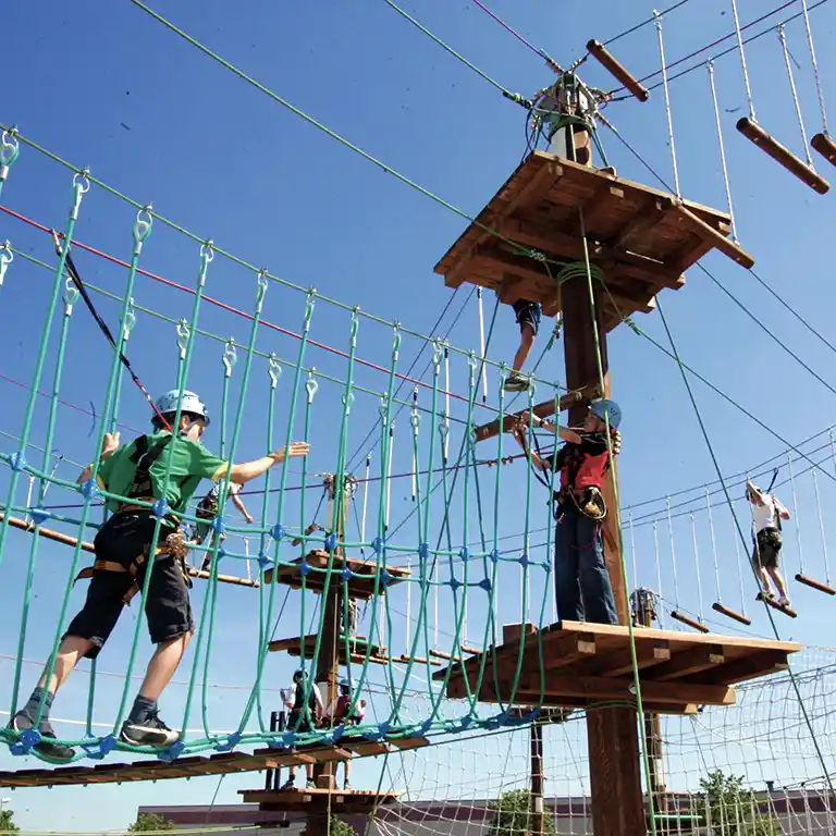 Several people wearing helmets and harnesses in the Südsee-Camp high ropes course, crossing rope bridges and wooden platforms under a blue sky.