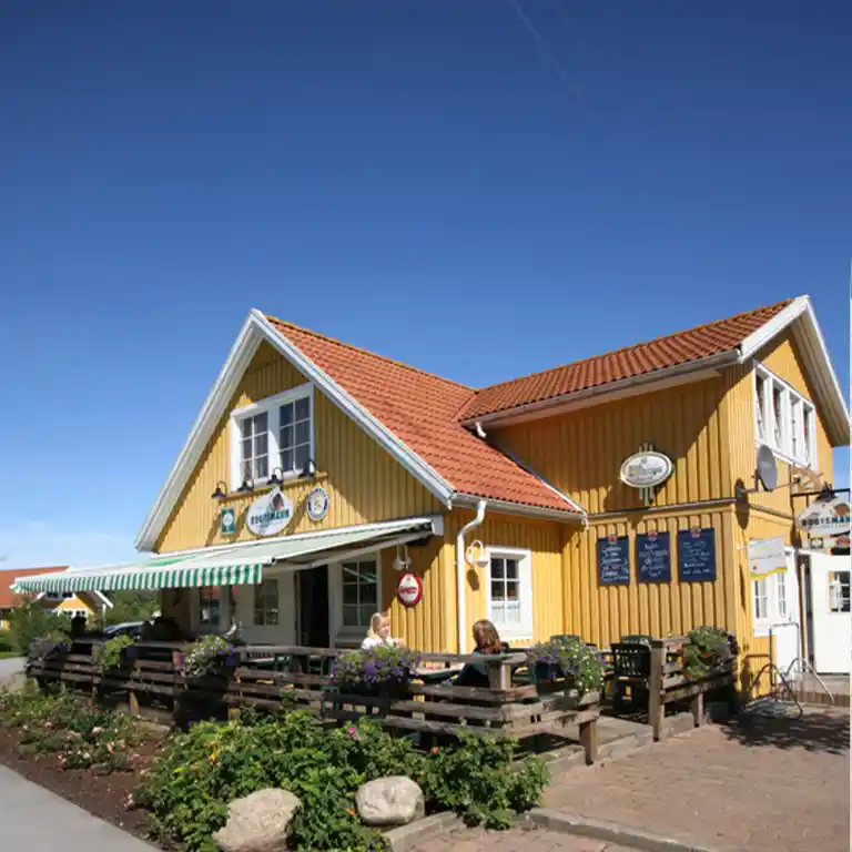 Scandinavian-style Bootsmann restaurant in the Sommarby holiday village at Südsee-Camp, with yellow wooden facade, red tiled roof, and outdoor seating with flower boxes.