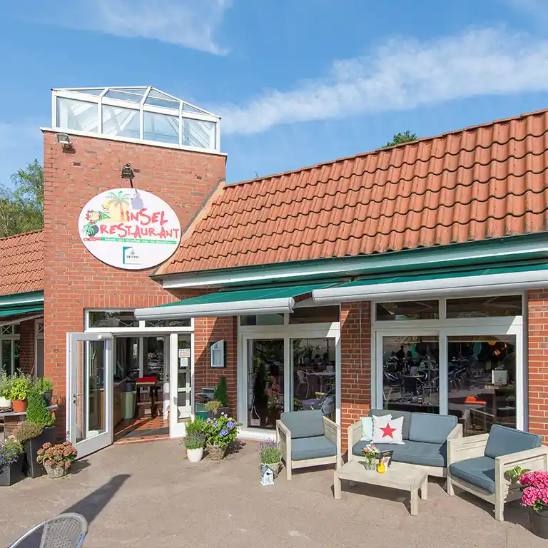 Entrance of the Insel-Restaurant at Südsee-Camp with red tiled roof, outdoor lounge seating, and open door to the terrace.