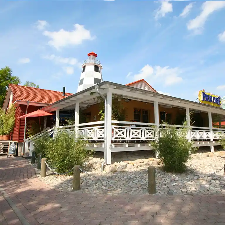 Pier One restaurant at Südsee-Camp with large veranda, maritime-style lighthouse, and white wooden facade under a blue sky.