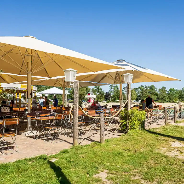 Beach bar at the lake in Südsee-Camp with wooden chairs under large umbrellas, located at the sandy shore with a view of the water and a red lighthouse.