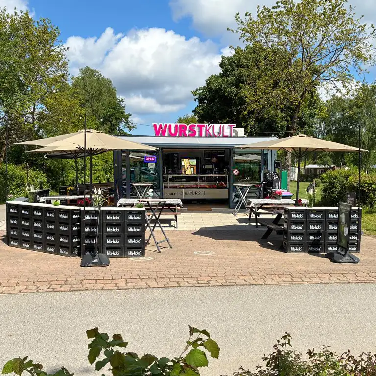 Snack stand at Südsee-Camp with the sign ‘Der Küstenhimmel wird jetzt zu WURSTKULT’ in front of a sausage counter, featuring the slogan ‘All you need is WURST’.