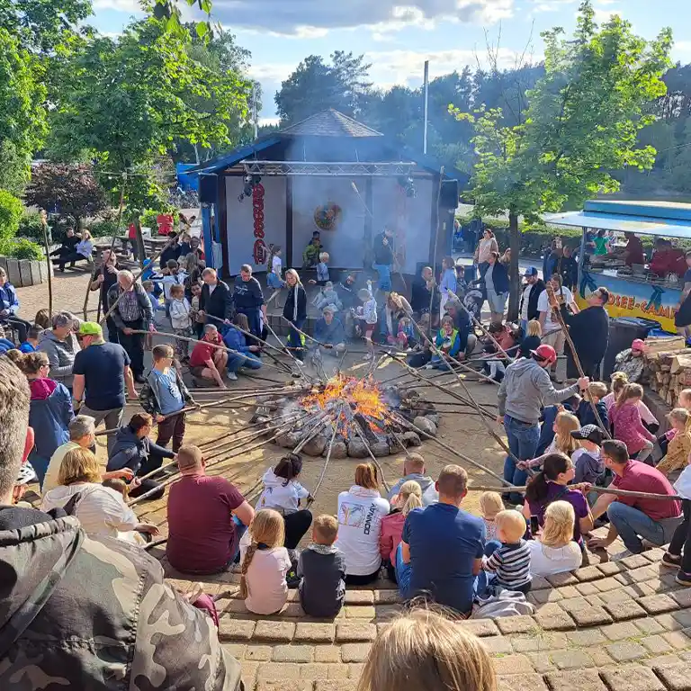 Many people of various ages sitting in a circle around a campfire, holding long sticks with dough pieces over the flames – enjoying communal bread baking at Südsee-Camp in a natural and cozy setting.