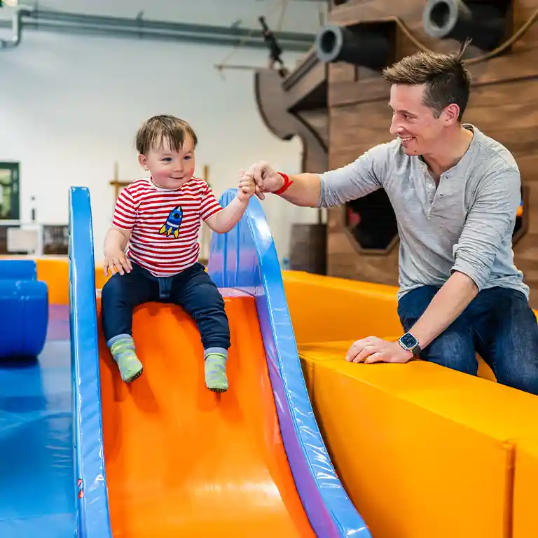 A father helps his toddler slide down in the indoor playground “Piratennest” at Südsee-Camp. The little child laughs and feels safe – a joyful moment for families with kids.