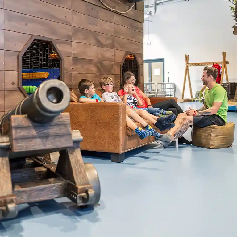 Children relax on a sofa at the indoor playground “Piratennest” at Südsee-Camp, while an adult chats and laughs with them. The pirate-themed setting with a cannon adds adventure – a place for both play and relaxation on a family holiday.