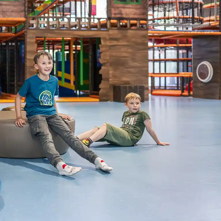 Two boys play in the indoor playground “Piratennest” at Südsee-Camp. They sit laughing on the floor in front of the large climbing and adventure area – a place full of fun for kids on a family holiday.