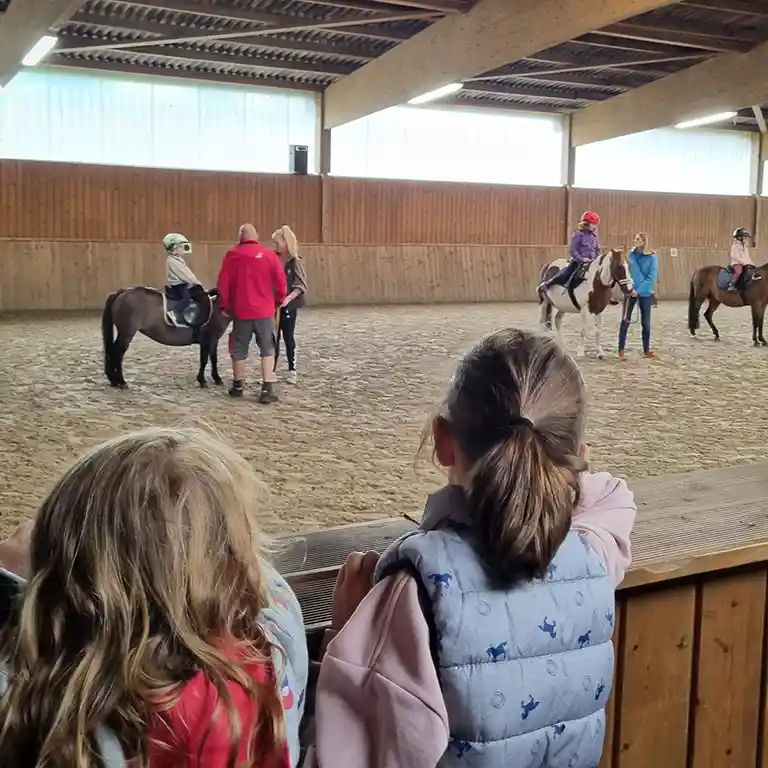 Children watch from the edge of the riding hall at Südsee-Camp as other kids ride ponies. Instructors provide guidance and ensure safety – an exciting holiday experience for families.