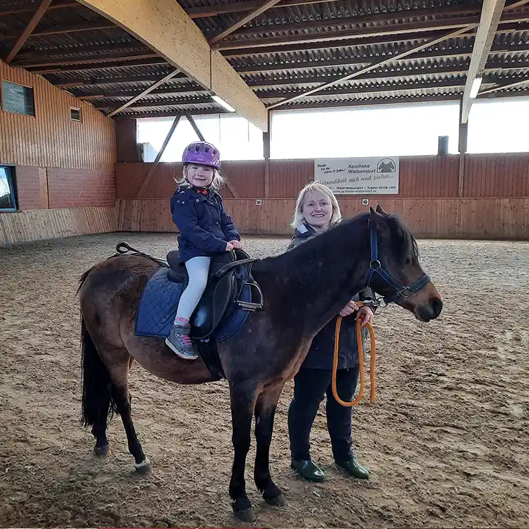 A little girl wearing a riding helmet sits happily on a pony in the riding hall at Südsee-Camp. An instructor holds the pony by the lead rope – a first riding experience for kids on a family holiday.