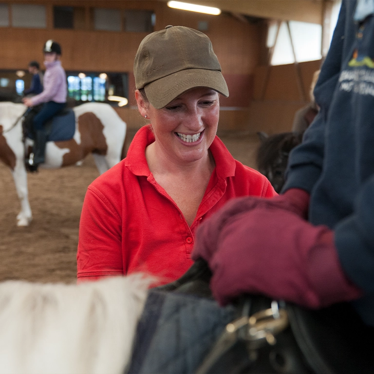 A riding instructor in a red shirt smiles during a lesson in the riding hall at Südsee-Camp. In the background, children are riding ponies – a joyful experience for families on holiday.