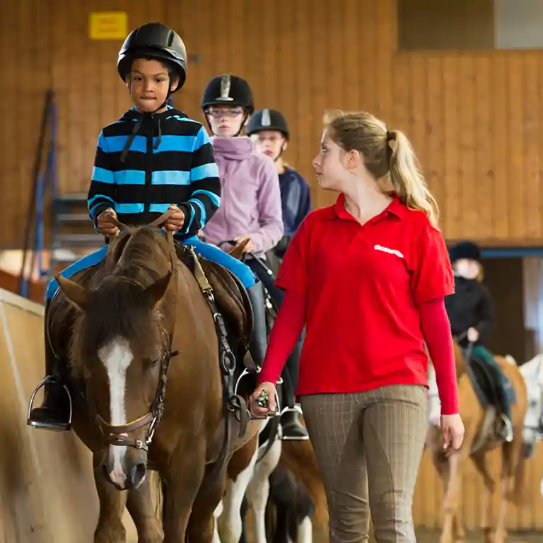 Children take part in a riding lesson at Südsee-Camp. A boy rides a horse while being guided by an instructor in a red shirt. Other kids follow on ponies inside the riding arena – an exciting holiday activity for families.