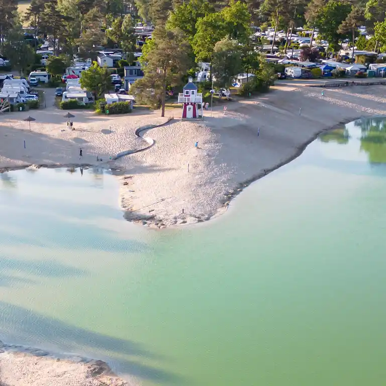 Luftaufnahme vom Sandstrand im Südsee-Camp mit Rutsche, kleinem Leuchtturm sowie Chalets und Mietwohnwagen direkt am Badesee.