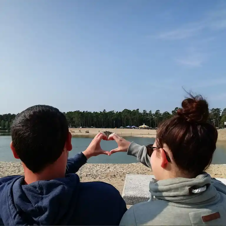 Ein Paar sitzt am Strand des Südsee-Camps und formt mit den Händen ein Herz mit Blick auf den Badesee und den Sandstrand. Ein romantischer Moment inmitten von Natur und Urlaubsatmosphäre.