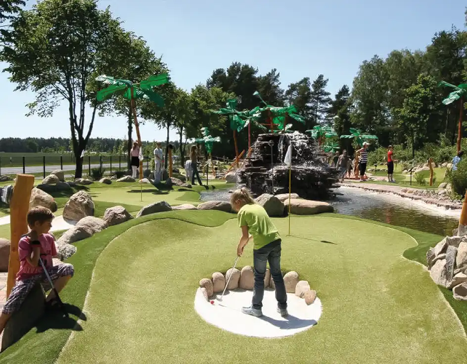 Children and families enjoy jungle mini golf at Südsee-Camp on a sunny day. The course is decorated with palm trees, rocks, and water features, creating a tropical adventure for all ages.