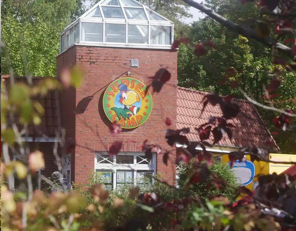 Brick building with the colorful Südsee-Camp logo – entrance to the “Nahkauf” supermarket at Südsee-Camp, surrounded by lush greenery.