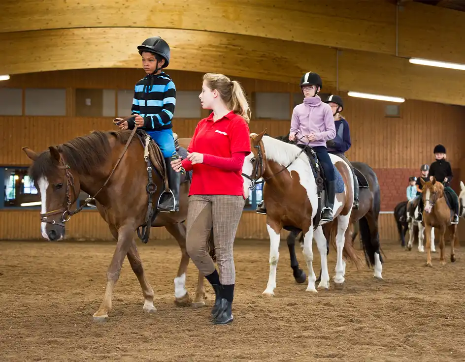 Children learning to ride horses at the Südsee-Camp riding school – indoor arena with instructor leading a riding lesson for beginners