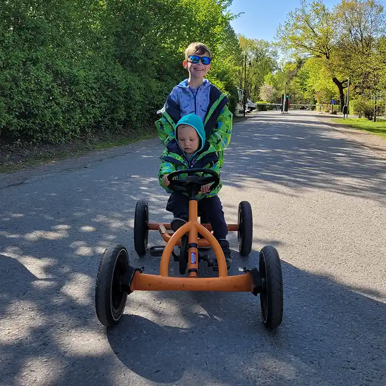 Two children at Südsee-Camp ride an orange go-kart on a paved road. The older boy stands behind the younger child and smiles while pushing – family fun in a holiday park.